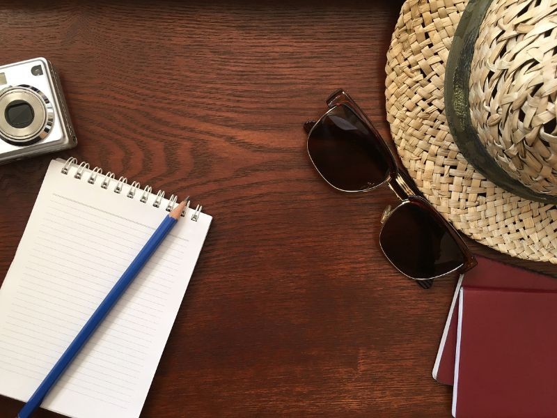 A notepad, sunglasses, camera and a sunhat on a wooden desk
